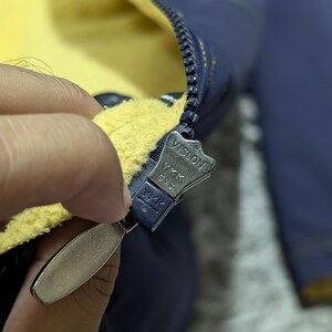 Może przedstawiać: Ciemnoniebieska kurtka z jaskrawoż&oacute;łtą podszewką z polaru. Kurtka posiada ciemnoniebieski plastikowy zamek błyskawiczny YKK Vislon z metalowym uchwytem. Suwak zamka jest wyraźnie oznaczony napisami Vislon, YKK oraz 5VS na swojej powierzchni.