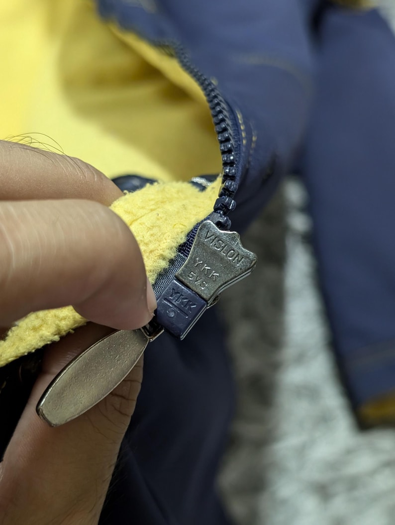 Może przedstawiać: Ciemnoniebieska kurtka z jaskrawoż&oacute;łtą podszewką z polaru. Kurtka posiada ciemnoniebieski plastikowy zamek błyskawiczny YKK Vislon z metalowym uchwytem. Suwak zamka jest wyraźnie oznaczony napisami Vislon, YKK oraz 5VS na swojej powierzchni.