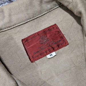 May include: A tan-coloured garment featuring a folded collar and a rectangular, dark red fabric label stitched onto the interior. The label displays an intricate, small emblem and the letter M on a separate white tag attached below the main label.