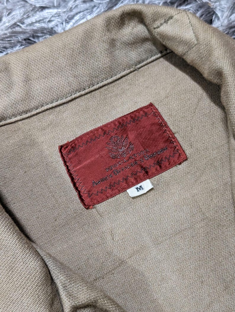 May include: A tan-coloured garment featuring a folded collar and a rectangular, dark red fabric label stitched onto the interior. The label displays an intricate, small emblem and the letter M on a separate white tag attached below the main label.