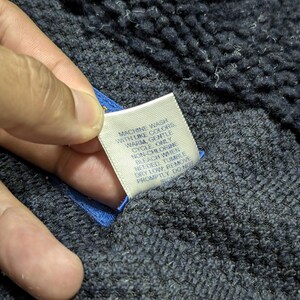 May include: Close-up of a navy blue textured fabric with a white tag attached. The tag has care instructions printed in black text: "MACHINE WASH WITH LIKE COLORS WARM, GENTLE CYCLE ONLY..." The fabric is held by a hand.