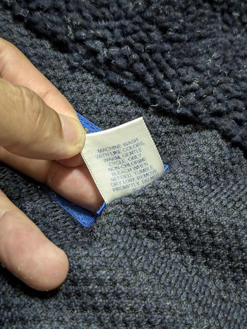 May include: Close-up of a navy blue textured fabric with a white tag attached. The tag has care instructions printed in black text: "MACHINE WASH WITH LIKE COLORS WARM, GENTLE CYCLE ONLY..." The fabric is held by a hand.