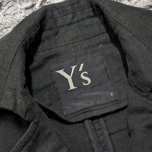 May include: A black blazer with a structured collar and a black fabric label sewn into the inner neckline. The label features the white text Y's and the number 2 printed in the bottom right corner. The garment has visible stitching along the seams.