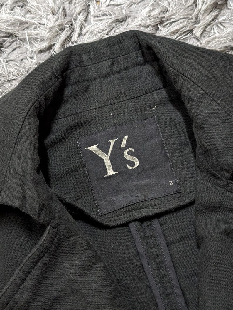 May include: A black blazer with a structured collar and a black fabric label sewn into the inner neckline. The label features the white text Y's and the number 2 printed in the bottom right corner. The garment has visible stitching along the seams.
