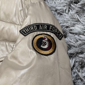 Peut inclure: Veste matelass&eacute;e beige avec une finition brillante. La veste pr&eacute;sente un &eacute;cusson sur la manche avec l'inscription "THIRD AIR FORCE" au-dessus d'un &eacute;cusson circulaire avec un motif "3".