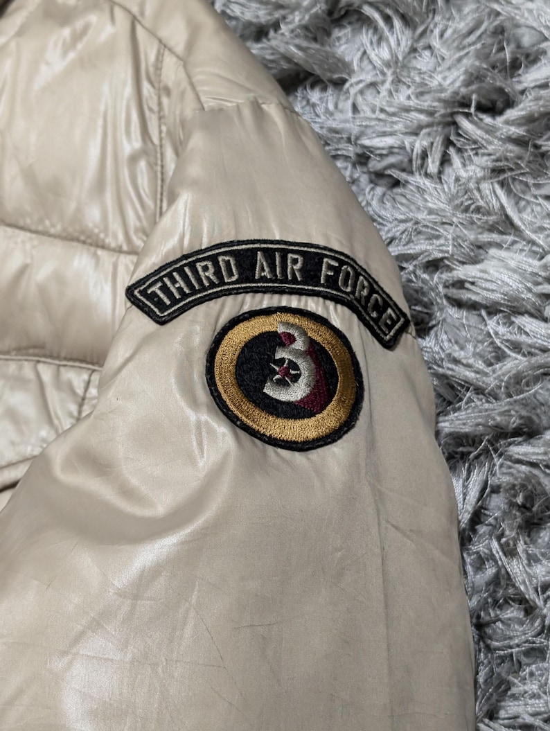 Peut inclure: Veste matelass&eacute;e beige avec une finition brillante. La veste pr&eacute;sente un &eacute;cusson sur la manche avec l'inscription "THIRD AIR FORCE" au-dessus d'un &eacute;cusson circulaire avec un motif "3".