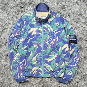 May include: A zip-up jacket featuring a multi-colored camouflage pattern in shades of blue, green, and cream. The jacket has a high collar, long sleeves with cuffs, and a black rectangular patch on the left sleeve with the text HH Helly Hansen SEA LIFE.