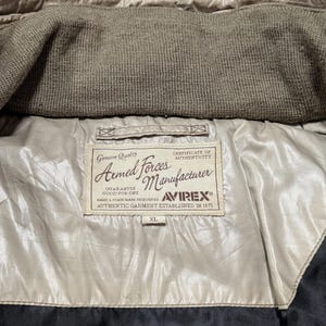 Peut inclure: Gros plan sur l'int&eacute;rieur d'une veste beige avec un col en tricot marron. La doublure de la veste est de couleur beige clair, et une &eacute;tiquette indique "Armed Forces Manufacturer" avec la marque Avirex et la taille XL.