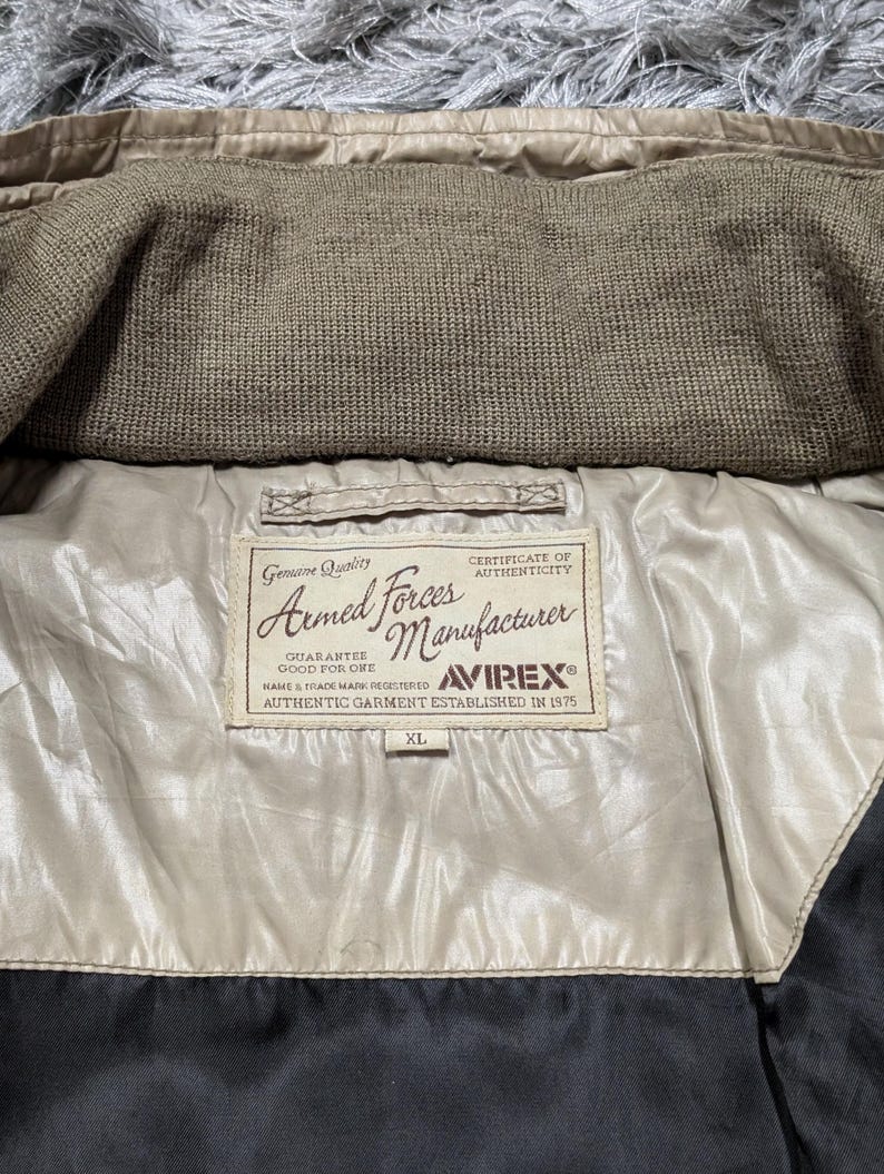 Peut inclure: Gros plan sur l'int&eacute;rieur d'une veste beige avec un col en tricot marron. La doublure de la veste est de couleur beige clair, et une &eacute;tiquette indique "Armed Forces Manufacturer" avec la marque Avirex et la taille XL.