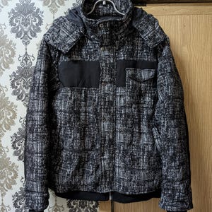 May include: A black and white textured jacket featuring a high collar, a detachable hood, and solid black panels across the chest. The garment includes a front button closure, a chest pocket with a flap, and black ribbed cuffs at the sleeves and hem.