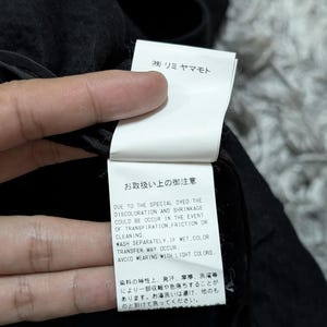May include: A white rectangular garment care label attached to a black fabric item. The label features black text in Japanese and English, including the brand name (株) リミヤマモト and instructions regarding potential discoloration, shrinkage, and color transfer.