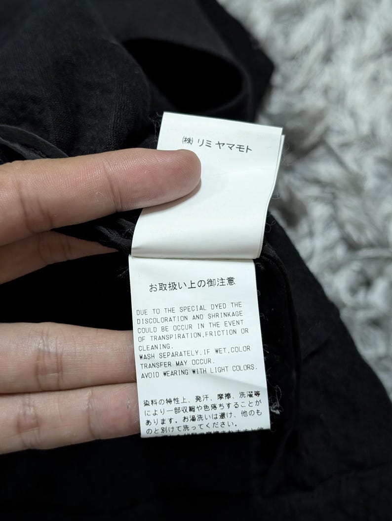 May include: A white rectangular garment care label attached to a black fabric item. The label features black text in Japanese and English, including the brand name (株) リミヤマモト and instructions regarding potential discoloration, shrinkage, and color transfer.
