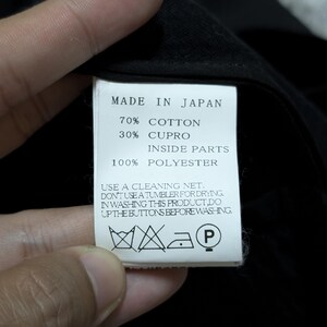 May include: A white rectangular garment care label with black text reading MADE IN JAPAN, 70% COTTON, 30% CUPRO, INSIDE PARTS 100% POLYESTER, and care instructions including USE A CLEANING NET, DON'T USE A TUMBLER FOR DRYING, and DO UP THE BUTTONS.