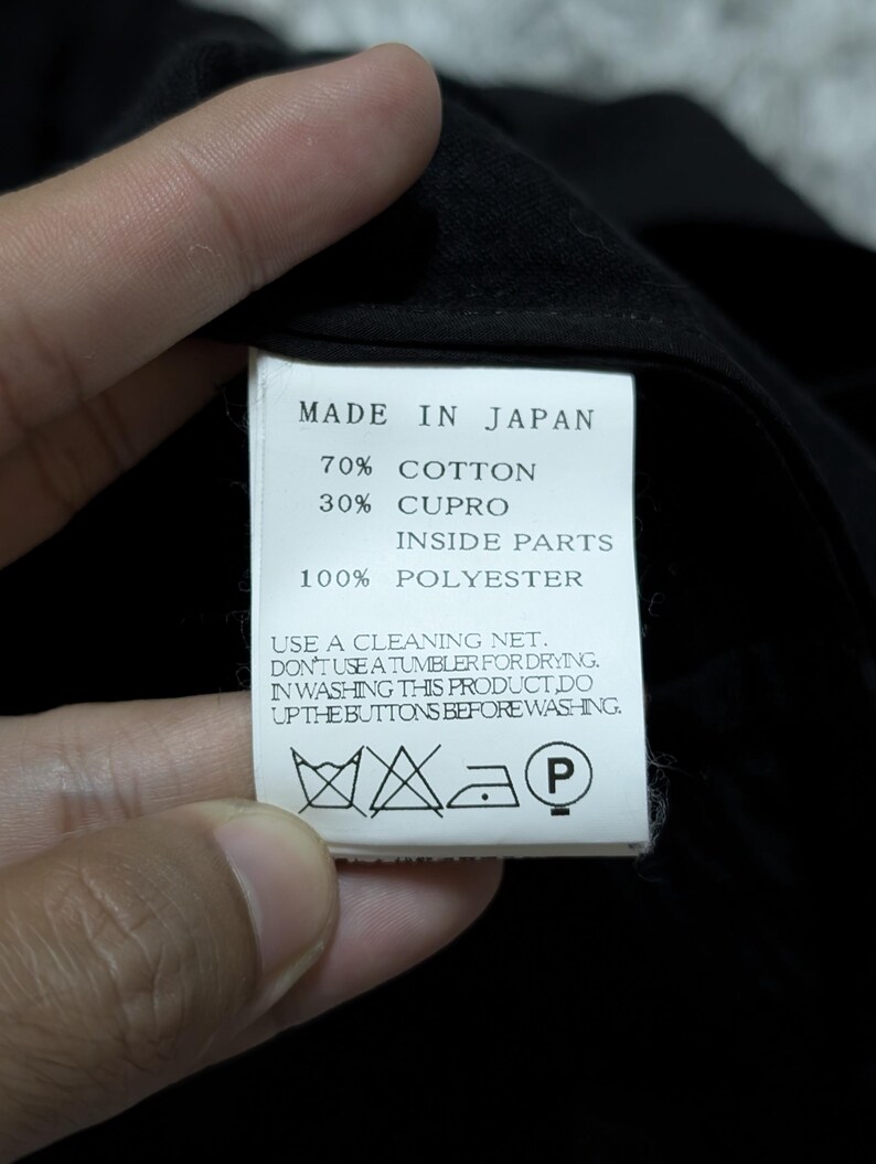 May include: A white rectangular garment care label with black text reading MADE IN JAPAN, 70% COTTON, 30% CUPRO, INSIDE PARTS 100% POLYESTER, and care instructions including USE A CLEANING NET, DON'T USE A TUMBLER FOR DRYING, and DO UP THE BUTTONS.