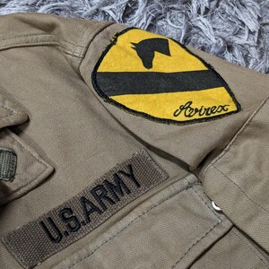 May include: A tan jacket featuring a rectangular patch with the text U.S. ARMY in black lettering. Above this, a shield-shaped patch displays a yellow background with a black horizontal stripe, a black horse head silhouette, and the word Avirex.