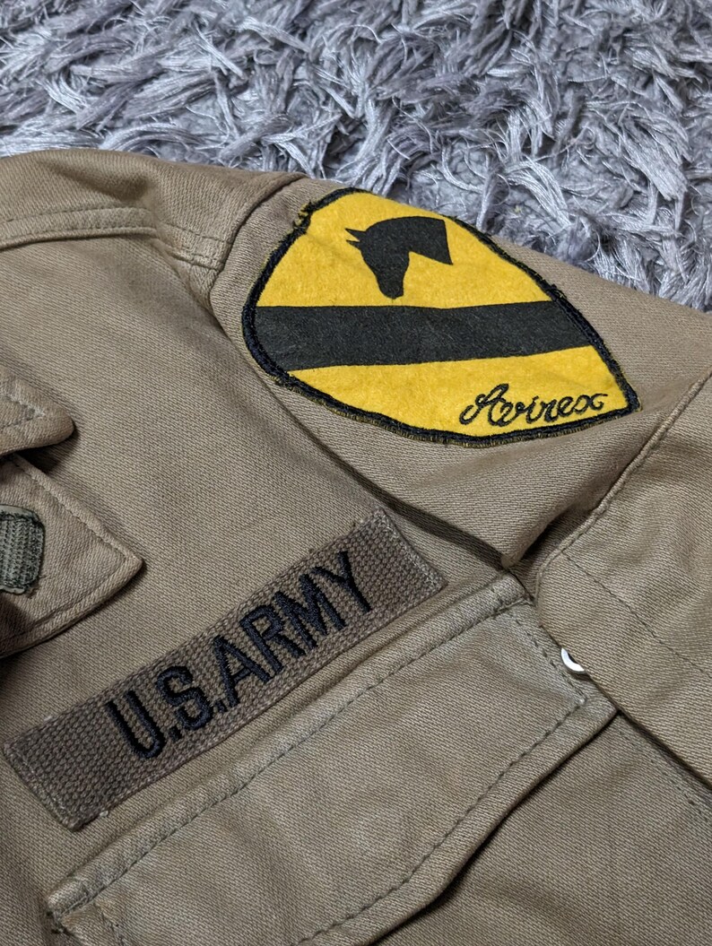 May include: A tan jacket featuring a rectangular patch with the text U.S. ARMY in black lettering. Above this, a shield-shaped patch displays a yellow background with a black horizontal stripe, a black horse head silhouette, and the word Avirex.