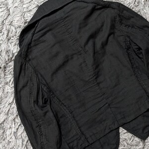 May include: A black jacket featuring a wide collar, long sleeves, and visible seam detailing along the shoulders and arms. The garment is constructed from a textured fabric with a slightly wrinkled appearance and a structured, tailored cut.