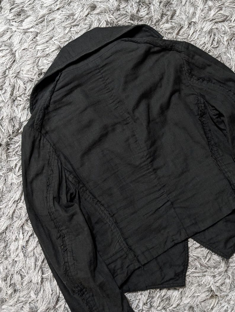 May include: A black jacket featuring a wide collar, long sleeves, and visible seam detailing along the shoulders and arms. The garment is constructed from a textured fabric with a slightly wrinkled appearance and a structured, tailored cut.