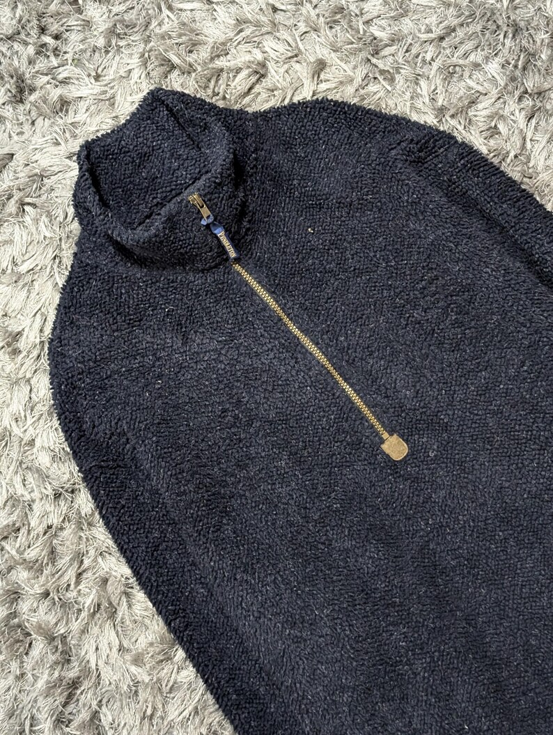 May include: A navy blue fleece pullover with a high collar and a gold-colored zipper. The textured fabric is visible in this close-up shot of the top. The zipper extends down the front of the garment.