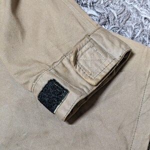 May include: A tan-coloured sleeve cuff made of a durable, textured fabric. The cuff features a rectangular tab with reinforced stitching and a small, black hook-and-loop fastener patch attached to the edge for adjustable closure and fit.