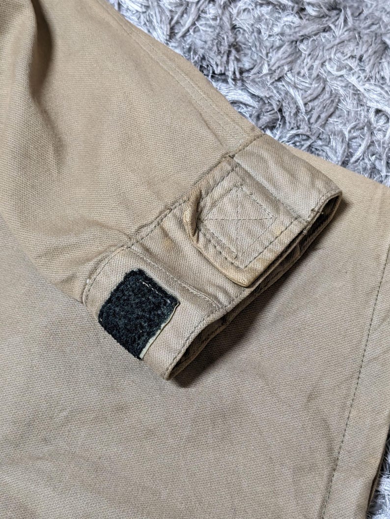 May include: A tan-coloured sleeve cuff made of a durable, textured fabric. The cuff features a rectangular tab with reinforced stitching and a small, black hook-and-loop fastener patch attached to the edge for adjustable closure and fit.