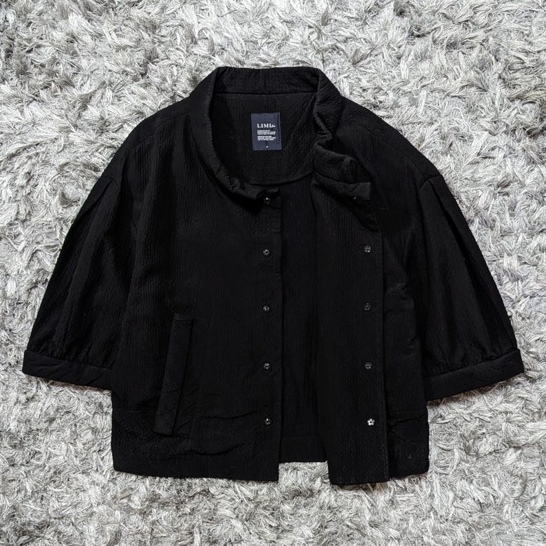 May include: A black button-down jacket with a textured fabric, featuring three-quarter length sleeves, a stand-up collar, and a front placket with visible buttons. A small rectangular label with the text LIMI feu is sewn inside the neckline.