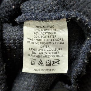 May include: Close-up of a navy blue knit fabric with a white tag. The tag includes text indicating the fabric composition: 70% acrylic, 30% polyester, and washing instructions. The tag also includes the text "ALSO SEE REVERSE".