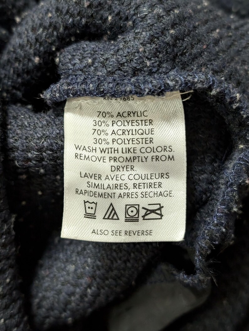 May include: Close-up of a navy blue knit fabric with a white tag. The tag includes text indicating the fabric composition: 70% acrylic, 30% polyester, and washing instructions. The tag also includes the text "ALSO SEE REVERSE".