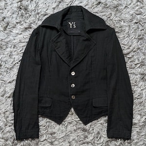 May include: A black blazer with a wide collar, three front buttons, and two small flap pockets at the waist. The interior label features the text Y's. The garment has long sleeves and a pointed hemline, constructed from a textured fabric.