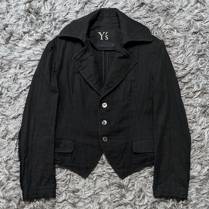 May include: A black blazer with a wide collar, three front buttons, and two small flap pockets at the waist. The interior label features the text Y's. The garment has long sleeves and a pointed hemline, constructed from a textured fabric.