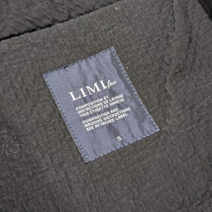 May include: A black garment with a textured, puckered fabric surface featuring a dark blue rectangular interior label. The label displays the text LIMI feu, followed by composition and washing instructions in French and English, with a size S at the bottom.