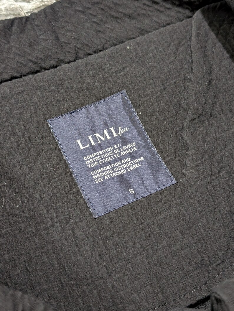 May include: A black garment with a textured, puckered fabric surface featuring a dark blue rectangular interior label. The label displays the text LIMI feu, followed by composition and washing instructions in French and English, with a size S at the bottom.