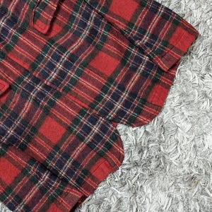 May include: Close-up of a red, green, and navy blue plaid fabric. The fabric is a classic tartan pattern, with a repeating grid of intersecting lines. The fabric appears to be a medium-weight material, possibly wool or a wool blend.