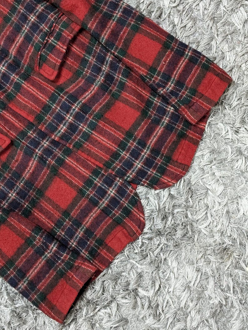 May include: Close-up of a red, green, and navy blue plaid fabric. The fabric is a classic tartan pattern, with a repeating grid of intersecting lines. The fabric appears to be a medium-weight material, possibly wool or a wool blend.
