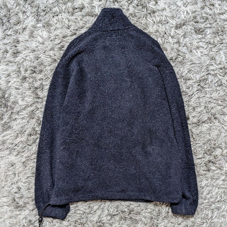May include: A navy blue fleece jacket with a high collar and a drawstring at the hem. The jacket has long sleeves and a soft, textured appearance. The jacket is displayed on a gray, textured surface.