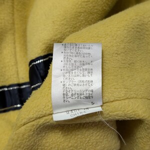 Może przedstawiać: Ż&oacute;łta odzież z polaru z białą metką z instrukcją prania w języku japońskim oraz napisem MADE IN CHINA. Po lewej stronie teksturowanej ż&oacute;łtej tkaniny widoczny jest ciemnogranatowy pasek ze srebrnymi metalicznymi nićmi.