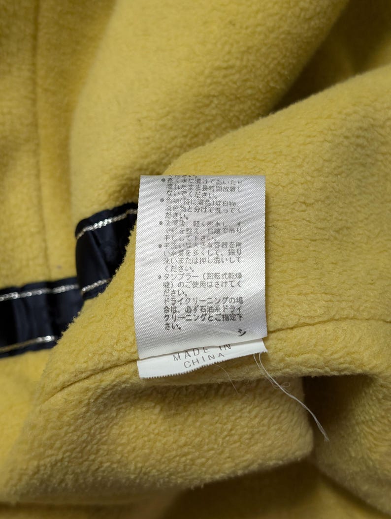 Może przedstawiać: Ż&oacute;łta odzież z polaru z białą metką z instrukcją prania w języku japońskim oraz napisem MADE IN CHINA. Po lewej stronie teksturowanej ż&oacute;łtej tkaniny widoczny jest ciemnogranatowy pasek ze srebrnymi metalicznymi nićmi.