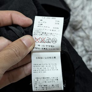 May include: A white rectangular garment care label attached to a black fabric item. The label features the model number LX-J15-005, material composition of 70% cotton and 30% cupro, with 100% polyester filling, alongside care instructions and warnings.