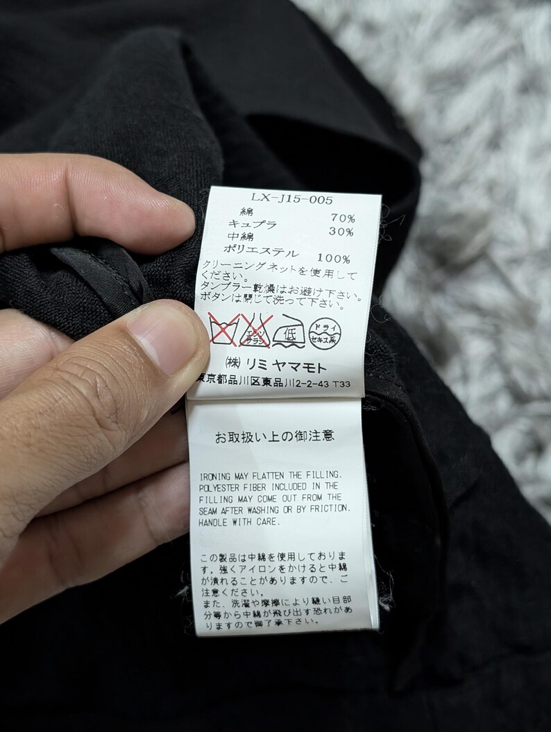 May include: A white rectangular garment care label attached to a black fabric item. The label features the model number LX-J15-005, material composition of 70% cotton and 30% cupro, with 100% polyester filling, alongside care instructions and warnings.