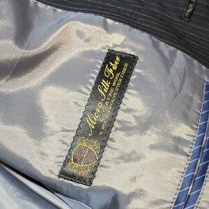 May include: Close-up of the interior lining of a dark pinstripe suit jacket. The lining is a light gray color, with a label that reads "Micro Silk Fiber" in gold lettering. The label also features a crest design.