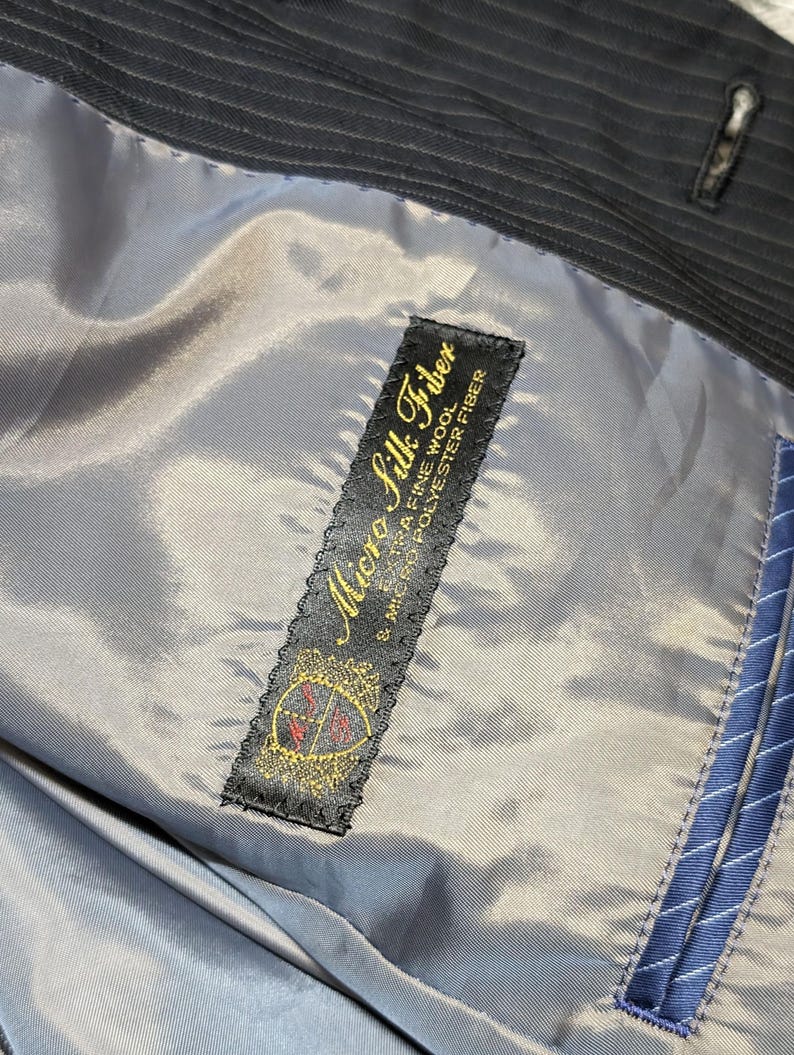 May include: Close-up of the interior lining of a dark pinstripe suit jacket. The lining is a light gray color, with a label that reads "Micro Silk Fiber" in gold lettering. The label also features a crest design.