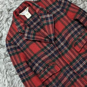 May include: A red, green, and navy plaid blazer with a notched collar and two front pockets. The blazer has a classic design and is made of a woven fabric. The label reads "Comme des Garcons".