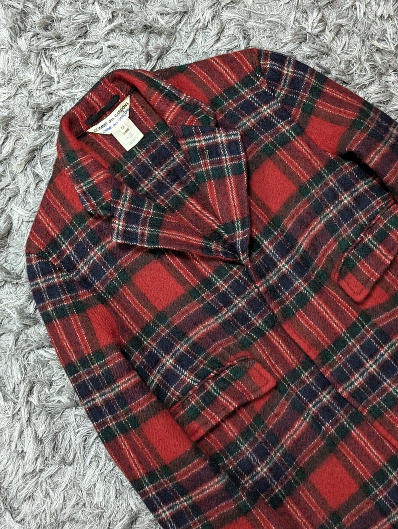 May include: A red, green, and navy plaid blazer with a notched collar and two front pockets. The blazer has a classic design and is made of a woven fabric. The label reads "Comme des Garcons".