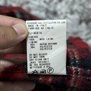 May include: A tag from Comme des Gar&ccedil;ons, made in Italy, with the text "CJ-40014". The tag details the fabric composition: 100% wool, 62% rayon, and 38% polyester. Care instructions include "Do Not Bleach" and "Dry Clean Only".