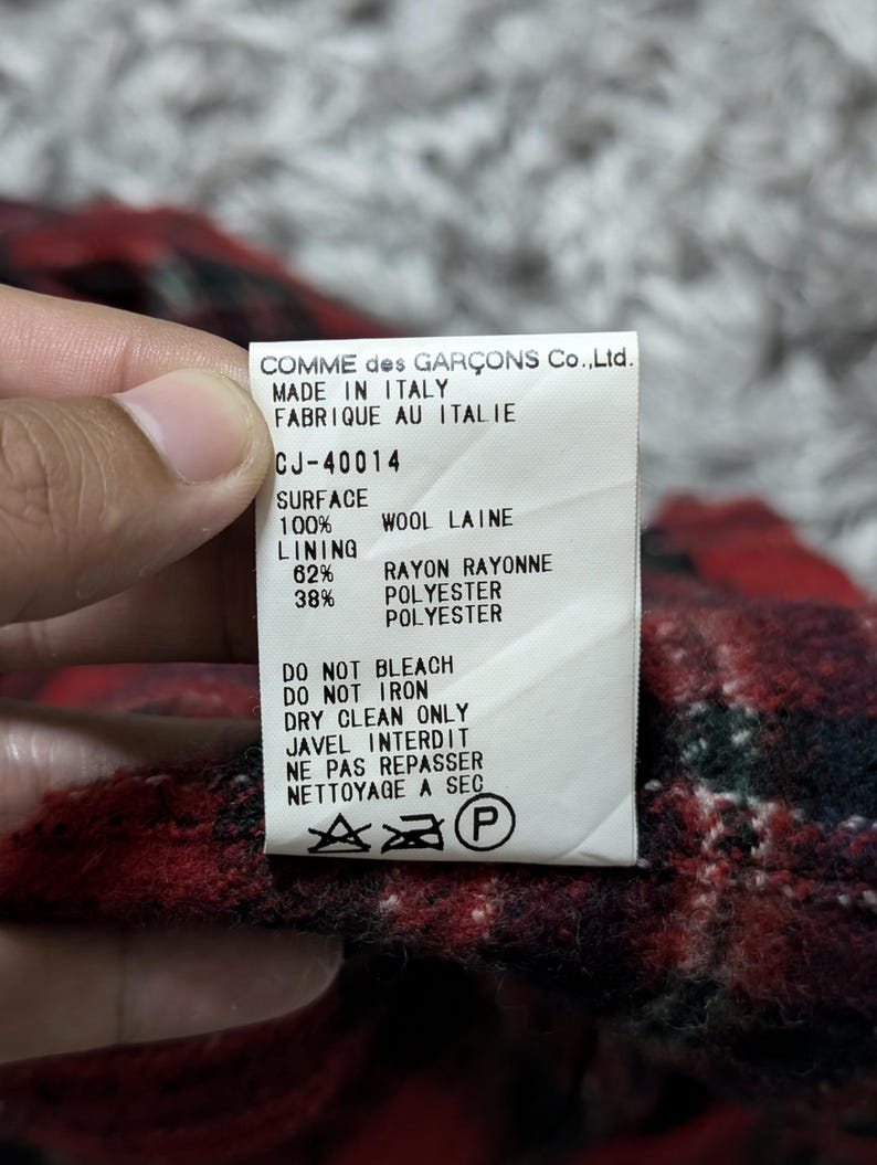 May include: A tag from Comme des Gar&ccedil;ons, made in Italy, with the text "CJ-40014". The tag details the fabric composition: 100% wool, 62% rayon, and 38% polyester. Care instructions include "Do Not Bleach" and "Dry Clean Only".