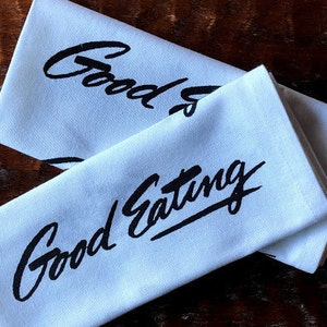 May include: Two white cloth napkins with black text. The top napkin says "Good" and the bottom napkin says "Good Eating".