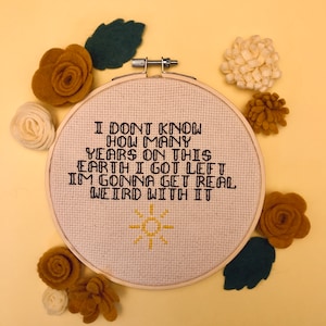 May include: A circular cross-stitch embroidery featuring the text "I DON'T KNOW HOW MANY YEARS ON THIS EARTH I GOT LEFT I'M GONNA GET REAL WEIRD WITH IT". The design includes a yellow sun. The hoop is surrounded by felt flowers and leaves.