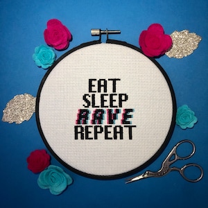 Puede incluir: Un aro de bordado blanco con letras negras que dicen "Eat Sleep Rave Repeat". El aro está rodeado de flores de fieltro de colores y purpurina.