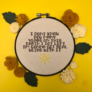 May include: A white embroidery hoop with a brown cross-stitch design that reads "I don't know how many years on this earth I got left I'm gonna get real weird with it". The hoop is surrounded by yellow felt flowers and green felt leaves on a yellow background.