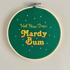 May include: A green embroidery hoop with white stars and the text "Well Now Then Mardy Bum" in yellow and pink lettering.